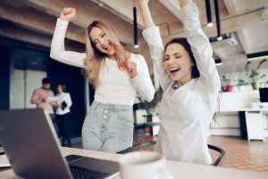 Two young happy businesswomen celebrating project success in modern office